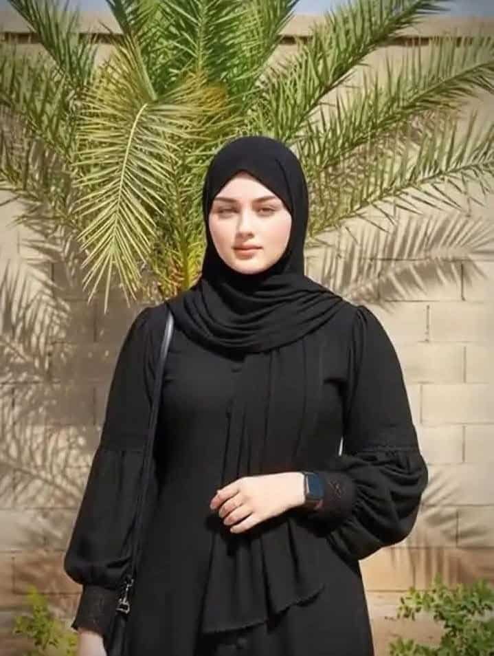 امرأة ترتدي عباءة سوداء وقف بجانب نخلة في الهواء الطلق.