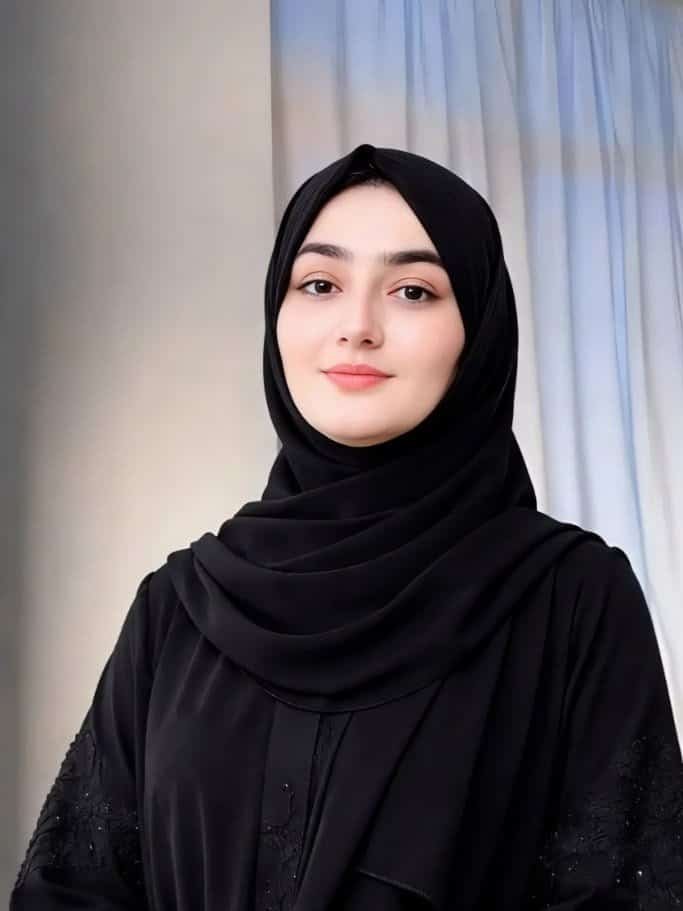 امرأة ترتدي حجاب أسود أمام ستارة بيضاء، صورة واضحة وهادئة.
